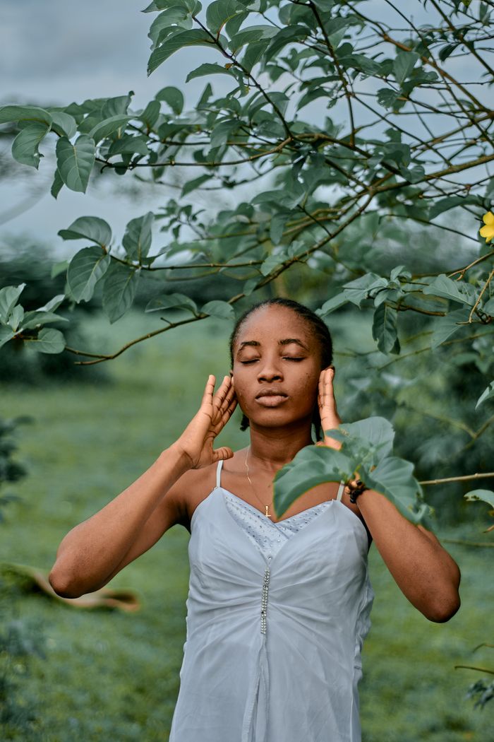 Portrait of a woman with eyes closed, surrounded by lush greenery in a peaceful outdoor setting.